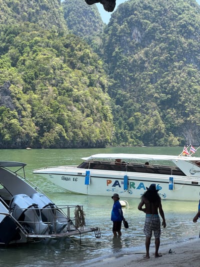 A photograph captures a coastal scene in Ban Ko Klang, Thailand. Dense green foliage covers the cliffs in the background. The water is a murky green color, where two boats are docked. In the foreground, part of a boat with four outboard engines is visible. Alongside a larger boat with the name "PATRASS" emblazoned on it, two men stand in the shallow water. One man is wearing a hat, a blue shirt, and shorts. The other man is wearing a hat and plaid shorts.
