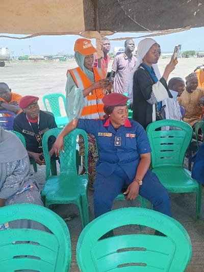 The image shows a group of people sitting on green plastic chairs under a canopy in Katabu, Nigeria. The coordinates, 10.71 latitude and 7.514 longitude, place the location in the Federal Capital Territory, Abuja, Nigeria. There are eight people present, including a woman in a blue uniform with a red beret, a woman in a green vest and a white headscarf, a man in a black robe and white headscarf, another woman in a black robe and white headscarf, and four other men. One man is holding a phone in his hand. The woman in blue is sitting in the foreground, looking at the camera. The woman in the green vest is looking down at her phone. The woman in the black robe is looking at the phone being held by the man next to her. The others are either looking straight ahead or at the camera.