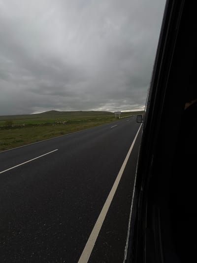 The image shows a view from inside a vehicle, likely a car, traveling on a road in Tlik, Armenia. The road is paved with dark asphalt and has painted white lane markings. To the left of the road is a grassy field with a hilly terrain. Above the field is a gray, overcast sky. Parts of the vehicle, including the frame and side mirror, are visible at the top of the image.