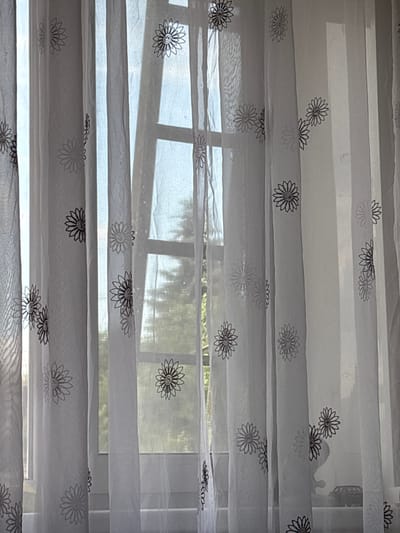 The image shows a close-up of sheer white curtains with a repeating floral pattern, hanging in front of a window. The window reveals a blurred view of trees and sunlight, suggesting an outdoor scene. The curtains are vertically pleated and appear to be made of a lightweight, translucent material. The floral pattern consists of stylized flowers with outlined petals in a dark color. The overall impression is one of domestic interior with a view to the outside. There is no discernible information in the image to indicate its location in Zhigulyovsk, Russia.