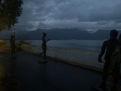 The image captures a nighttime scene on the lakeside promenade in Montreux, Switzerland. The foreground features dark, wet pavement reflecting minimal light. A single streetlamp with a yellowish glow is visible on the far left, partially illuminating a tree and the path. Three dark, bronze-colored human-shaped statues are positioned along the promenade. From left to right, the first statue is a standing female figure, partially obscured by the streetlamp. The second statue is a female figure in a dynamic pose, appearing to step forward or dance, with one leg raised and arms extended. The third statue, on the right, depicts a male figure carrying a large string instrument, possibly a cello, at his side. A light-colored, decorative metal balustrade separates the promenade from the expansive, dark waters of Lake Geneva. Across the lake, faint, scattered lights delineate a distant shoreline. The background consists of large, dark mountains whose peaks are partially obscured by heavy, low-hanging clouds under an overcast sky.