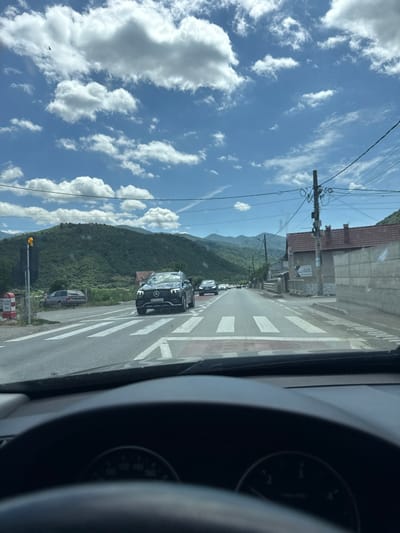 The image captures a view from inside a car driving on a two-lane road in Boița, Romania. The road is marked with white pedestrian crossings, and several cars are visible, including a black Mercedes-Benz SUV in the foreground, traveling in the same direction. The car is approaching a crosswalk. To the left of the road, there is a traffic light, a utility pole with wires, and a parked older model car. Further in the distance, houses with red-tiled roofs and a hill covered in lush green vegetation can be seen. The sky is blue with scattered white clouds, and a bird is flying in the upper left quadrant of the image. The dashboard of the car is partially visible in the foreground, showing the speedometer.