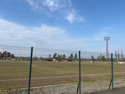 The image displays an outdoor sports field, identified as being in the city of ქალაქი ლანჩხუთი, Georgia. The perspective is from behind a dark green wire mesh fence that occupies the lower half of the frame, positioned above a gravel-covered ground strip. The field itself is a grass pitch, marked with white lines, indicating its use for a sport like football (soccer), and appears somewhat worn in areas.
On the field, approximately twenty individuals are visible. Some wear light-colored jerseys (appearing white or light blue), while others are in darker attire. They are distributed across the pitch, suggesting engagement in a game or practice session, though specific actions are indistinct due to distance. Along the far perimeter of the field, a series of advertising banners or signs are visible. A digital scoreboard is situated in the mid-ground, towards the center-right of the field.
A prominent, tall, rust-colored metal floodlight tower with a rectangular light fixture stands on the right side of the background, beyond the field. The background also includes a line of trees, some with sparse, autumnal foliage, bordering the playing area. The sky above is clear blue with scattered white, wispy clouds.