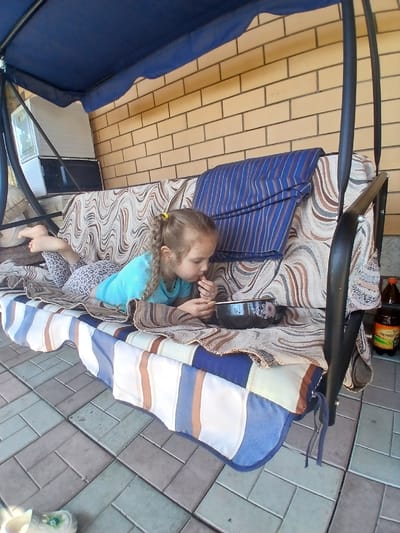 A young girl with blonde hair in a ponytail, wearing a blue t-shirt and leopard-print leggings, lies on a swing set's upholstered seat, eating from a black bowl. The swing is positioned outdoors, in front of a light brown brick wall. A blue canopy covers the top of the swing. The ground is paved with interlocking gray and green tiles. To the right of the swing, on the ground, are two bottles, one of which has a label with Russian text.