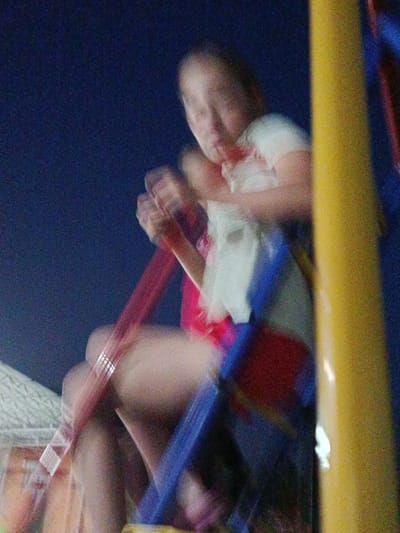 An outdoor nighttime image shows a person on a swing set. The background is dark blue, suggesting a night sky. The person appears to be young, with light-colored hair, and is wearing a white t-shirt with red accents. They are holding onto the red chains of the swing. The swing set structure is yellow and blue. The image is blurry due to motion. The location is specified as Novotitarovskaya, Russia.