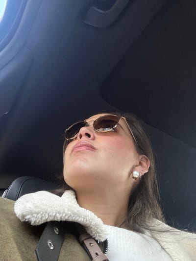 A low-angle shot of a woman wearing sunglasses and a white pearl earring. She is positioned in the lower-right corner of the frame with a dark gray ceiling in the background. She wears a light brown jacket with a white faux fur collar. A seatbelt with black and silver hardware is visible. Her lips are slightly parted, and her skin has a dewy finish. The sunglasses are round with gold frames and brown lenses.