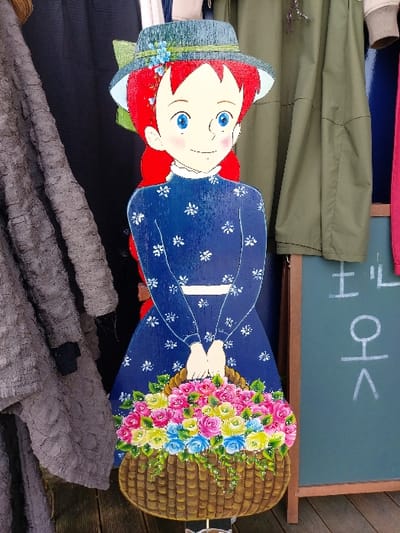 A two-dimensional, life-sized wooden cutout of a female character with red hair, blue eyes, and rosy cheeks stands centrally. The character wears a blue dress with white floral patterns, a green straw hat adorned with blue flowers, and holds a brown woven basket overflowing with pink, yellow, and blue roses. To the cutout's left, a textured gray fabric, possibly a knitted garment, hangs beside a dark fabric backdrop. To its right, olive-green fabric drapes next to a dark blue surface. Further right, a dark green blackboard with light brown wooden framing displays white Korean characters ("토", "오", "ㅅ"). The cutout rests on a wooden plank surface, indicating an outdoor or semi-outdoor retail setting, likely a storefront or market stall in 강화군, South Korea.
