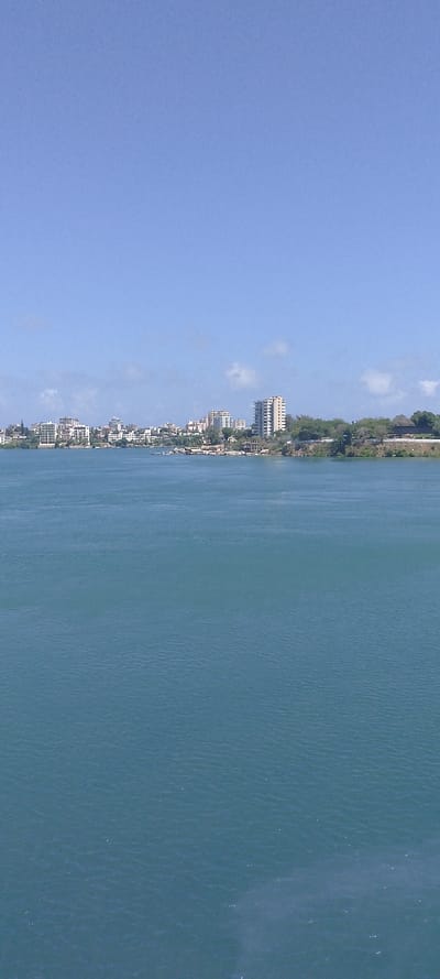 The scene depicts a wide, elevated view across a large body of water towards the urbanized coastline of Tudor ward, Kenya, under a clear, light blue sky with minimal, scattered white clouds near the horizon. The lower two-thirds of the image are dominated by expansive, calm blue-green water exhibiting subtle surface ripples. The distant shoreline, spanning the central horizon, is characterized by a dense arrangement of multi-story buildings, predominantly light-colored, interspersed with green foliage. Several taller high-rise structures are visible, particularly towards the right side of the urban cluster. No individuals, active vessels, or specific actions are discernible within the frame.