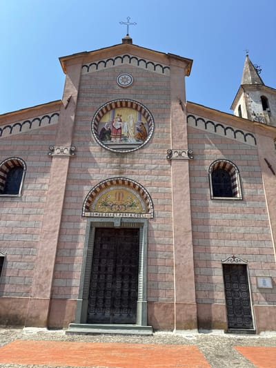 The image shows the facade of a church in Apricale, Italy. The church has a pink and gray brick exterior with decorative elements. There is a large central entrance with ornate double doors, flanked by two smaller doors. Above the main entrance is a mosaic depicting a religious scene. A bell tower with a clock face is visible to the right of the church. The sky is clear and blue.