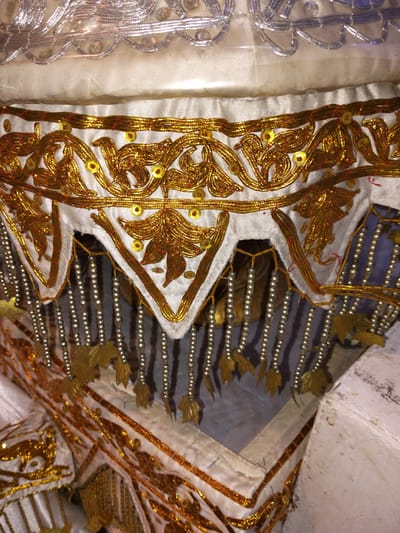 This image displays a close-up view of an ornate textile, likely part of a ceremonial garment or altar decoration. The fabric is primarily white and embellished with intricate gold embroidery featuring swirling patterns and leaf-like motifs. Small yellow sequins are scattered across the embroidered sections, adding sparkle. Dangling from the lower edge of the textile are strands of small, silver-colored beads, interspersed with small, metallic gold leaf-shaped charms. A silver, wavy trim runs along the top edge of the fabric, also appearing to be made of metallic thread. The background is a muted blue, suggesting the textile is displayed against a colored surface. No people or specific actions are visible; the focus is entirely on the detailed craftsmanship of the decorative textile. The location in Cottrueng, Indonesia, is not visually evident from the image itself.