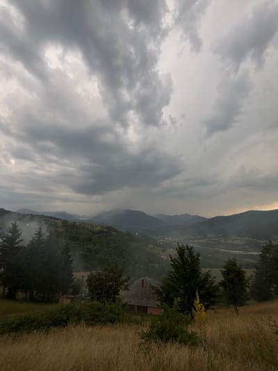 The image depicts a panoramic view of a rural mountainous landscape under a heavy, cloudy sky, suggesting approaching or ongoing rainfall. In the foreground, dry, tall grass covers a hillside. A small, weathered building with a red roof and a darker-colored lower section is situated on this hillside, partially obscured by dense green foliage and trees. To the left of this building, several tall, dark green coniferous trees are visible, with a plume of white smoke rising from behind them, indicating a possible fire or burning activity.
The midground is characterized by rolling hills covered in dense green forests, with a scattering of smaller buildings and homes visible throughout the valleys and on the slopes. A road or river can be faintly discerned in the distance. The background features a range of layered mountains fading into a hazy, muted atmosphere. The sky is filled with dramatic, dark gray clouds, with lighter patches suggesting breaks in the cloud cover or distant sunlight. A distinct vertical streak of lighter gray, possibly rain, is visible in the far distance over the mountains.
The scene appears to be a remote, natural environment. The location, identified as Mekića Kula, Montenegro, suggests a mountainous region with a rustic, possibly agricultural or sparsely populated setting. The overall atmosphere is somber and dramatic, influenced by the overcast weather conditions. No people are visible in the frame.