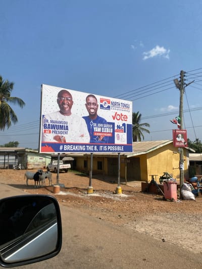 This outdoor shot, taken in Hohoe, Ghana, captures a political campaign billboard along a rural or semi-urban roadside under a clear blue sky. The image appears to be taken from a vehicle, with a rearview mirror visible in the bottom right corner.

The central subject is a large, brightly lit billboard featuring two smiling male politicians. The top figure is identified as "DR. MAHAMUDU BAWUMIA AS PRESIDENT," wearing glasses and a light-colored shirt. Below him is "DR. JOHN SAVIOUR YAW ELEBLU AS MEMBER OF PARLIAMENT," dressed in a blue and white patterned shirt. The billboard also displays "NORTH TONGU Constituency," "Vote No. 1," and the hashtag "#BREAKING THE 8, IT IS POSSIBLE," indicating an ongoing election campaign.

In the background, there's a dusty roadside and a paved road. A power pole with overhead wires stands tall, and a smaller red and white banner, possibly for another candidate named "MAHAMA," is affixed to it. Further back, a yellow/ochre-colored building with a dark roof is visible. A black and white cow stands near the base of the billboard on the right side. The scene is brightly lit by the sun, suggesting daytime with clear weather. The overall impression is a bustling roadside in Ghana during an election period.
