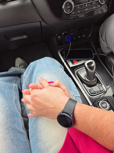 In the interior of a car, two people hold hands over light-wash denim jeans. One person wears a black smartwatch on their left wrist and a magenta-colored jacket. The other person has pink nail polish. A smartphone lies next to the gear shift. The car’s dashboard is visible, including climate control buttons and the start/stop engine button.
I do not have the information to verify the location of the city in Аур, Russia.