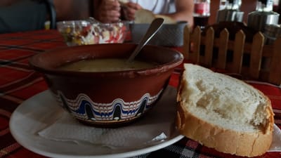 An overhead shot shows a traditional Bulgarian meal, likely in the city of Pevtsite, Bulgaria, as indicated by the food and decor. The meal consists of a bowl of soup, a plate of salad, and a slice of bread. The soup is served in a brown clay bowl with blue and white decorative patterns. A spoon rests in the soup. The salad is in a clear glass bowl and appears to contain tomatoes, lettuce, and corn. A slice of white bread sits to the right of the soup bowl. The table is covered with a red and black checkered tablecloth. In the background, a small wooden fence and two glass bottles are visible. A person's hands can be seen reaching for bread in the background. The lighting is natural and suggests daytime.