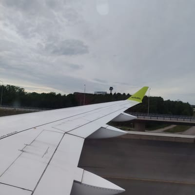 The image displays a forensic view from an aircraft window, revealing the white wing of an AirBaltic aircraft parked on an asphalt apron at Stockholm-Arlanda Airport, Sweden. The wing's upper surface, marked with "NO STEP OUT" text and various panel lines, occupies the lower two-thirds of the frame. The wingtip features a distinctive lime-green section with "airBaltic" printed in white. Beyond the wing, a dark green treeline separates the immediate airport ground from the background elements. Prominently positioned above the treeline, a multi-story air traffic control tower with a distinctive, multi-tiered cabin is visible to the right. To the left of the control tower, a large, light-colored airport building stands. A highway bridge with multiple lanes crosses horizontally in the midground, partially concealed by the foliage. The sky is uniformly overcast and grey, with a soft, diffused light, and a subtle circular lens flare appears in the upper right quadrant. No people are visible.