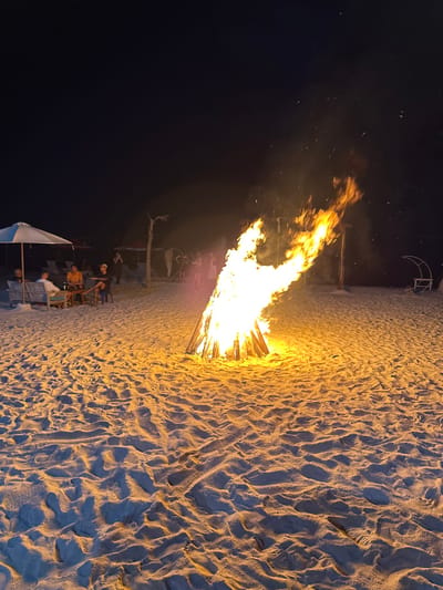 The image depicts a nighttime scene on a sandy beach in Jungutbatu, Indonesia. A large, conical bonfire, composed of stacked logs, burns intensely in the mid-ground, emitting bright orange and yellow flames and ascending sparks. The fire illuminates the surrounding sandy terrain, which shows numerous footprints and surface irregularities. To the left, under a white beach parasol, three individuals are seated on wooden benches around a low, square wooden table. One person wears a white shirt, another an orange shirt, and a third a dark top. Further in the background, to the right of the seated group, indistinct figures are visible standing and mingling. Notable background objects include a heart-shaped archway constructed from natural materials, situated slightly left of the bonfire, and a white, crescent moon-shaped structure to the far right. Additional beach furniture and structures are dimly visible in the shadowed distance against the dark sky.