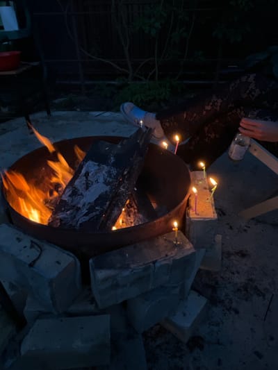 In Вельяминово, Russia, at night, a close-up shot captures a fire pit filled with burning logs, casting an orange glow. Several small, lit candles are placed on bricks surrounding the pit. A person's legs, wearing a blue shoe, are visible on the left. On the right, another person's hand holds a glass bottle. The scene appears to be an outdoor gathering, possibly a celebration, with the fire and candles creating a warm, intimate atmosphere despite the darkness of the surroundings.