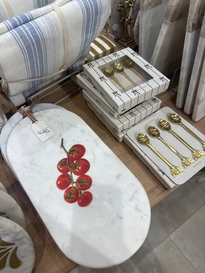 This image, captured in Milnerton, South Africa, showcases a display of kitchenware or home goods. Dominating the scene are several stacks of boxed gold-colored spoons, some topped with butterfly-shaped embellishments. Adjacent to these boxes is a white marble serving board adorned with a printed tomato design. A price tag is attached to the board. Additionally, a stack of blue-and-white striped towels is visible on the left side. The arrangement suggests a retail environment.