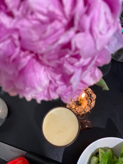 In Podgorodnyaya Pokrovka, Russia, a still life is depicted on a dark, flat surface. The foreground is dominated by a large, out-of-focus pink flower, likely a peony, obscuring much of the scene. To the right of the flower, a small, lit candle glows in a decorative, gold-colored holder adorned with clear, faceted stones. In front of the candle, a dark brown mug filled with a light-colored beverage, possibly coffee or tea with milk, is placed. Partially visible to the left is a portion of a clear glass object, and to the far left, a silver knife with a black handle. In the lower right corner, a white bowl contains a salad, with visible pieces of cucumber and leafy greens. The lighting suggests an indoor setting, possibly at dusk or in a dimly lit room, with the candle providing a warm, localized illumination.