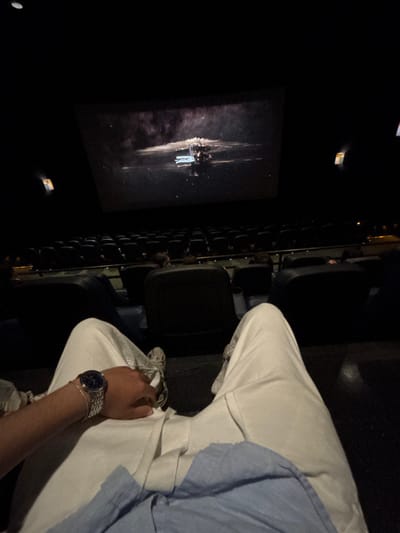 The image captures a first-person perspective from inside a dark cinema hall, showing a movie playing on the screen and a partially visible person. The individual, likely seated in an aisle seat, is wearing white or light-colored pants and a light blue collared shirt. Their right arm is visible, resting on their lap, adorned with a silver-toned watch with a dark face and a thin bracelet.
To the left of the person, several rows of dark, unoccupied cinema seats are visible, extending into the background. The low lighting typical of a movie theater creates a dark ambiance, with only the screen and minimal ambient light illuminating the space.
On the large screen, a science fiction scene is displayed. It features a prominent dark structure or spaceship with intricate details and some bright elements, possibly reflecting light or being part of the vessel's design, set against a dark, starry, or cosmic background. The image suggests a quiet and immersive movie-watching experience. There is no visible text.