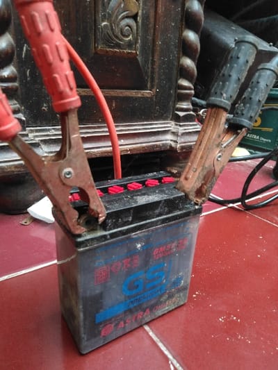 A used GS Astra Premium GM5Z-3B 12V5Ah lead-acid motorcycle battery, black with blue and white labeling, sits on a red tiled floor with white grout lines. Two jumper cable clamps are attached to its terminals: a red-handled clamp to the left terminal (presumed positive) with a red cable extending rearward, and a black-handled clamp to the right terminal (presumed negative) with a black cable extending to the right. The battery's top features six red caps covering the cell vents. The exterior of the battery shows signs of use and minor corrosion. Behind the battery, a dark, carved wooden furniture piece with spiral and floral motifs is visible. To the right rear, a dark green object, possibly another battery or power unit labeled "ASTRA," is partially discernible. Black cables are visible in the background on the right side. The red tiled floor has scattered white particulate matter on its surface. The scene is located in Mornington, Australia.