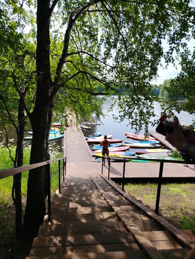 The image depicts a wooden staircase leading down to a wooden pier on a lake. The pier is lined with numerous colorful paddleboards. A shirtless man stands on the pier, facing away from the camera. In the background, other people are visible on the pier and in the distance. Trees with green foliage overhang the scene, casting shadows on the stairs and pier. The lake appears calm and reflects the sky. The location is identified as the village of Vylytdor, Russia.