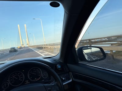 The photograph shows a driver's view from inside a car traveling across the Queen Elizabeth II Bridge, part of the Dartford Crossing in Dartford, United Kingdom. The bridge's support towers are visible through the windshield, as are other vehicles on the roadway. The dashboard instruments are visible, indicating a speed of approximately 40 mph. The side mirror shows a glimpse of the road and bridge structure behind the car. The date and time displayed on the dashboard are 30.05.26 at 6:03 AM with a temperature of +17.0°C.