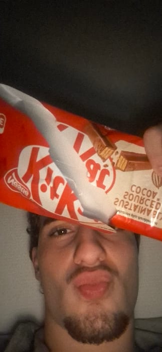 A person holds a Kit Kat bar, torn open, close to their face. The Kit Kat wrapper is red and white with the brand name prominently displayed. The person has dark hair, light skin, and is looking towards the camera. The background appears to be a dark or dimly lit space. The image is taken at an angle, giving a distorted perspective.