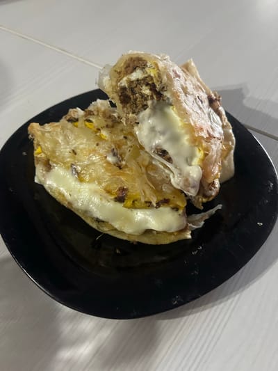 This media file shows a close-up, overhead view of a plate of food. The plate is black and round. On the plate are two pieces of a folded, flaky pastry filled with what appears to be ground meat and melted cheese. The cheese is gooey and is spilling out from the pastry. The pastry itself is golden brown and appears to be cooked. The background is a white surface with faint vertical lines, possibly a table. The lighting suggests it might be indoors, with artificial light casting shadows. There are no people or text visible in the image. The context provided indicates the location is Noyabrsk, Russia.