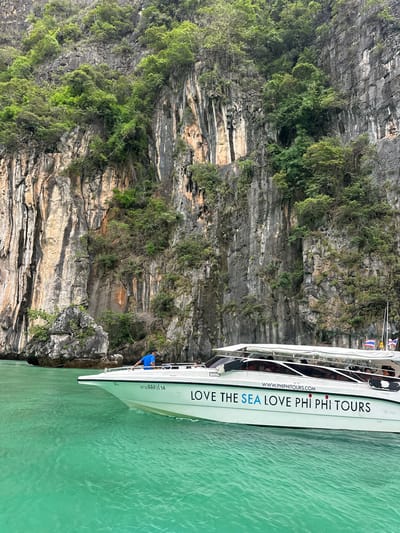 A white speed boat named "LOVE THE SEA LOVE PHI PHI TOURS" cruises through turquoise waters near a towering limestone cliff face covered in lush green vegetation. A person in a blue shirt stands near the front of the boat, leaning over the railing. Several passengers are visible inside the boat's cabin. Thai flags are attached to the boat's canopy. The cliff face is characterized by its rugged texture, vertical striations, and pockets of green foliage growing from its crevices. The water exhibits a vibrant, translucent green hue, with gentle ripples on its surface. The scene suggests a tourist excursion in a tropical, coastal environment.