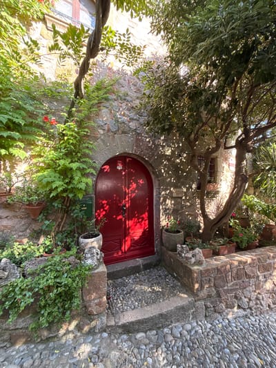 This is a photograph of a charming, sun-dappled scene in Tossa de Mar, Spain. The focal point is a rustic stone building with a vibrant red, arched doorway. The door features decorative paneling and a visible lock mechanism. To the left of the door, a lush green vine with small red flowers is climbing the stone wall, adding a splash of color.
Above and to the left of the doorway, a large tree with an old, gnarled trunk and dense green foliage casts dappled shadows across the stone wall and the ground. The sunlight filters through the leaves, creating a beautiful play of light and shadow. Several potted plants are strategically placed around the entrance, including a large, ornate pot to the right of the door with a trailing plant.
The building itself is constructed from rough-hewn stone, characteristic of older European architecture. To the right of the doorway, the path is paved with smooth, grey pebbles, a common feature in Mediterranean coastal towns. A small, green mailbox is mounted on the wall to the left of the door.
In the upper portion of the image, a window with a dark frame is visible within the stone wall, suggesting the interior of the building. The overall atmosphere is tranquil and inviting, evoking a sense of old-world charm and a peaceful Mediterranean afternoon. There is a small red sticker or label on the red door, but the text is not legible. The scene appears to be set during daylight, likely late morning or early afternoon, given the strong sunlight and distinct shadows.