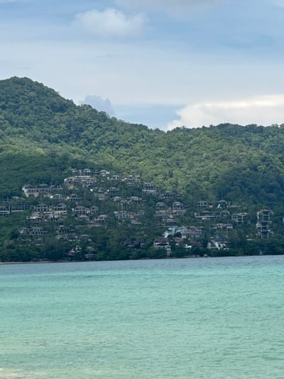 The image depicts a scenic view of a lush green mountain slope dotted with numerous buildings, cascading down towards a tranquil, turquoise sea. The buildings appear to be modern villas or resort accommodations, integrated into the natural landscape. The dense foliage of the tropical mountain covers much of the structures, creating a harmonious blend of architecture and nature.
The sea exhibits gentle ripples, suggesting calm weather conditions. The water's clarity allows for a glimpse of the seabed in some areas, further enhancing the idyllic tropical setting. The sky above is partly cloudy, with patches of blue sky interspersed with white clouds. A bright, luminous band of light, possibly a sunbeam or a reflection, cuts vertically through the sky on the left side of the image.
The scene is likely set in the late afternoon or early evening, given the soft lighting. The location appears to be a popular tourist destination, possibly Ban Bang Thao in Thailand, known for its beautiful beaches and resorts nestled amidst hills. There is no visible text in the image. The overall mood conveyed is one of serenity, luxury, and natural beauty.