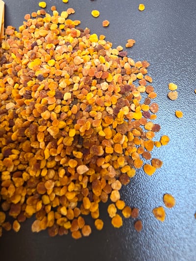 The image shows a pile of bee pollen granules on a dark, textured surface. The granules are small, irregular in shape, and vary in color from bright yellow to orange and reddish-brown. Some granules are rounder, while others are more elongated. A portion of a yellow object, possibly packaging, is visible in the upper left corner. The background is dark and slightly reflective. The scene appears to be an indoor close-up shot, with the lighting emphasizing the texture and color of the bee pollen. No specific location in Indzhe Voyvoda, Bulgaria, is depicted or suggested by the image content.