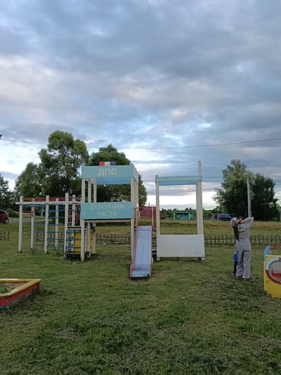 In the city of Smolino, Russia, a playground is depicted under a cloudy sky. A woman in gray athletic wear stands with her hands behind her head, facing away from the camera, near a playground structure. To her left, a child in blue clothing stands by the same structure. The playground features a monkey bar setup with blue and white bars, a yellow slide, and a sandpit with a red and yellow border. Signs are visible on the playground structures, one reading "ДПС" and another "ДЕЖУРНАЯ ЧАСТЬ," and on a separate structure, "ШКОЛА ДОРОЖНОГО ДВИЖЕНИЯ." In the background, trees line the horizon, and a dark-colored car is parked on a road. The ground is covered in grass, with some areas appearing recently mowed.