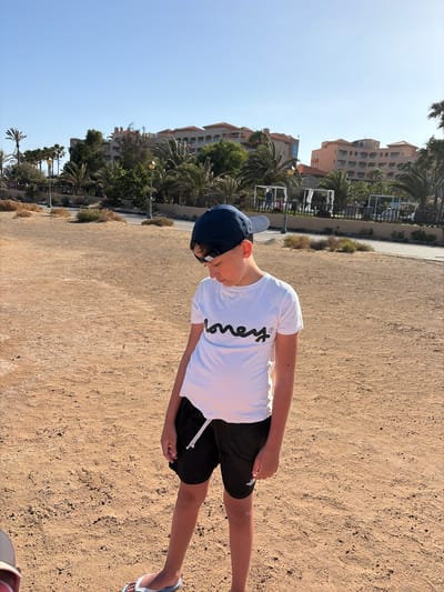 This image captures a young boy standing on dry, light brown sandy ground under a clear blue sky. The scene is located in Antigua, Spain. The boy, appearing to be pre-teen or early teen, is dressed in a white short-sleeved t-shirt with the black script "Honey®" printed on the chest. He wears black athletic shorts with a white drawstring, a dark blue baseball cap worn backward with sunglasses resting on its brim, and white flip-flops with light blue straps. He is looking downwards and slightly to his left.

Behind the boy, a paved road or path runs horizontally, bordered by a low fence and sparse, dry bushes. In the background, numerous palm trees and other green foliage frame multiple multi-story resort-style buildings. These buildings feature light-colored facades and red-tiled roofs. Several white canopy structures, possibly cabanas or outdoor seating, are visible near the buildings, with indistinct figures of people present in the distant resort area. The entire scene is brightly illuminated by direct sunlight, indicative of a clear day.