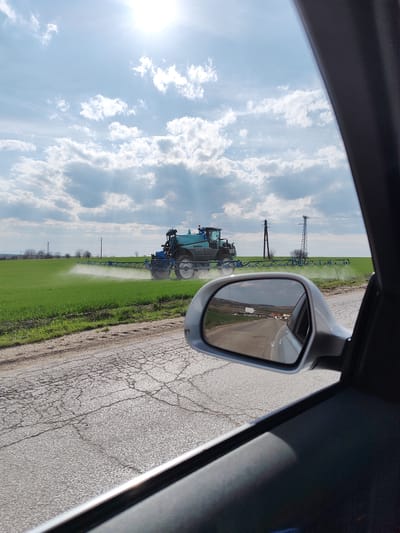 The scene is observed from the interior of a vehicle in the vicinity of Sofronievo, Bulgaria, looking forward and to the left. A teal and blue self-propelled agricultural sprayer, identified by visible text as a Berthoud model, is actively operating in a large, cultivated green field. The sprayer's wide, horizontal booms are extended, discharging a fine white spray onto the crops. The foreground features a cracked asphalt road adjacent to the field. In the mid-ground, several utility poles with power lines are visible, extending towards a distant, low-lying horizon. The sky is partly cloudy, displaying patches of blue amidst cumulus clouds, with bright sunlight emanating from the upper right, casting visible rays. The silver-colored passenger side mirror of the viewing vehicle occupies the lower right quadrant, reflecting the cracked asphalt road receding behind, along with distant hills and a small cluster of buildings. No individuals are visible in the immediate scene or the reflection.