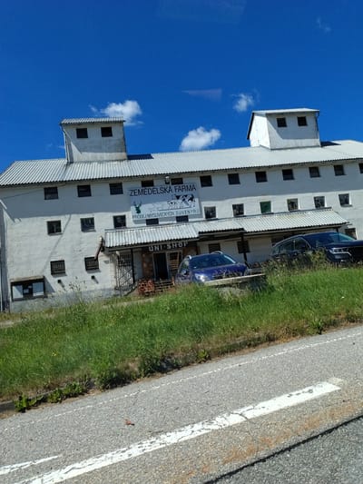 The image shows a white, multi-story building with a corrugated metal roof, identified by a sign as "ZEMEDELSKA FARMA" (Agricultural Farm). The sign also indicates "PRODEJ MYSLIVECKYCH SUVENYRŮ" (Sale of Hunting Souvenirs). The building has numerous windows and two distinct tower-like structures on the roof. In front of the building, there is a covered area with a sign that reads "UNI-SHOP." Two cars are parked in front of the shop, a blue hatchback and a dark SUV. The building is situated alongside a grassy embankment and a paved road. The sky is bright blue with a few scattered clouds. The image appears to have been taken from a vehicle moving along the road.