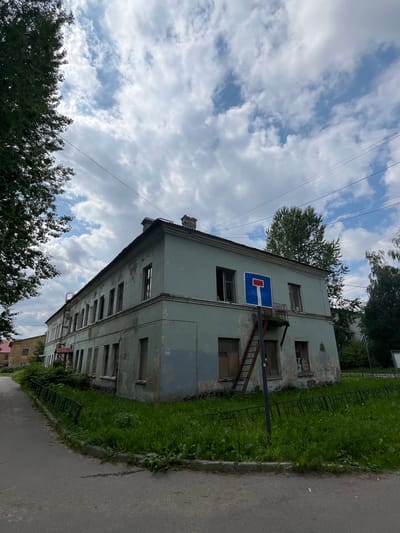 An overcast sky with scattered white clouds hangs above a weathered, light blue, two-story building located in the city of Imeni Sverdlova, Russia. The building, which appears to be an apartment complex or an older municipal building, exhibits peeling paint and some broken windows. A fire escape, made of rusted metal, is attached to the side of the building. A blue and red "dead end" street sign is mounted on a pole in front of the building. The building is surrounded by overgrown green grass and a low, black metal fence. To the left of the building, a paved road curves away from the viewer. The overall scene depicts an urban environment with signs of aging and disrepair.