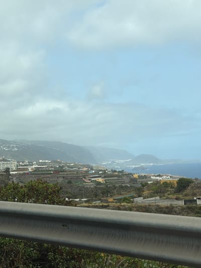 The image depicts a panoramic view of a coastal city in Spain. In the foreground, a metal guardrail runs horizontally across the frame, with some greenery and bushes visible below it. Beyond the guardrail, a terraced landscape unfolds, featuring buildings, agricultural fields, and sparse vegetation. The mid-ground reveals a denser urban area with various structures, including houses and commercial buildings, extending towards the coastline. In the background, a mountain range rises, partially shrouded in mist or clouds. The sea is visible on the right side of the image, with a clear horizon. The sky is overcast with clouds. The overall scene suggests a partly cloudy day with a view overlooking a town nestled between mountains and the sea.