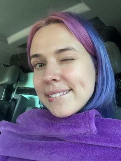The image is a close-up, slightly angled selfie of a smiling woman with heterochromatic hair, colored purple and blue, who is winking her right eye. She is wearing a purple, textured garment. The setting appears to be inside a car based on the visible headrest and interior features. The location within the city of Le Muy, France, cannot be determined from the visual data. No specific actions or interactions can be discerned beyond the subject taking a selfie.