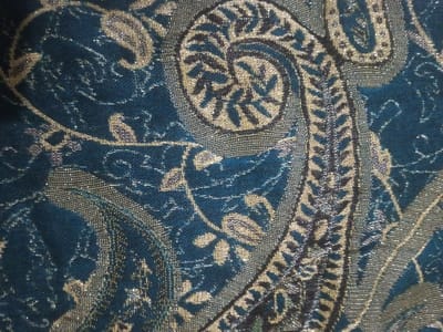 A close-up shot reveals a textile with a detailed paisley pattern. The dominant color is a deep teal, overlaid with intricate designs in metallic gold and silver threads. The paisley motifs feature swirling shapes with leaf-like elements within their borders and scattered around. The overall texture appears rich and potentially textured, suggesting a high-quality or ornate fabric.