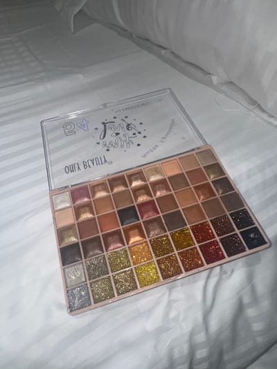 The image shows an open eyeshadow palette with a variety of colors, including shades of brown, gold, and glitter. The palette is on a white surface, possibly a bed or other soft material. The lid of the palette is clear with lettering. No people are visible. The environment suggests an indoor location, likely a bedroom. The location in the city of Gromshin, Bulgaria is not discernable from the visual information provided.