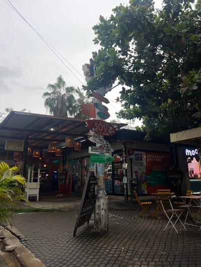 The image depicts a street scene in El Tunco, El Salvador, characterized by a cluster of businesses and directional signage. A prominent multi-directional signpost with various arrow-shaped signs indicating destinations such as "SOY LOFTS," "GINGER HOUSE," and "VERALUNA SPA" stands near the center. Beneath the signs, storefronts are visible, including "CUSCATLAN ATM" and "ROAD MARKET." Outdoor seating with tables and chairs is arranged on a paved walkway in front of some establishments. Lush green foliage from trees and a palm tree are present, suggesting a tropical environment. The sky is overcast. There are no visible people actively interacting, though some merchandise is displayed outside shops. A dog is lying down in the background.