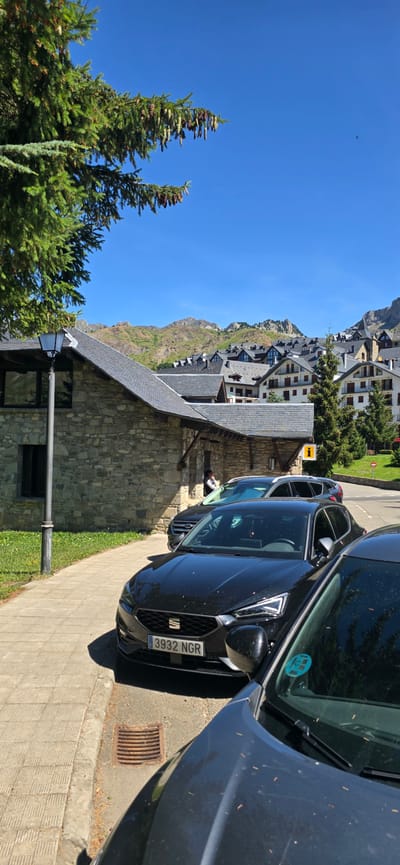 This is a vertical, eye-level, outdoor, daytime photograph taken in Sallent de Gállego, Spain. The foreground features the hoods of two black cars parked on a paved surface next to a sidewalk. The license plate of the closer car reads "3932 NGR". Behind the cars, a stone building with a slate roof is visible, with a lamppost on its left. Further back, a town with dense buildings and steeply pitched roofs stretches across a hillside, leading up to mountainous terrain under a clear blue sky. The architecture of the town suggests it is a ski resort. A few evergreen trees are scattered throughout the scene.