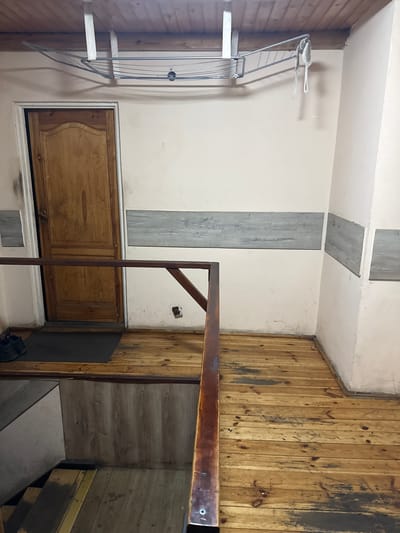 This is an indoor scene, likely a hallway or landing area, in Wąwelnica, Poland. The flooring is made of unfinished wooden planks with visible wear and some dark staining. A dark brown wooden railing runs diagonally across the foreground, obscuring a staircase descending into a lower level which has wood-paneled walls. To the left, a wooden door with a curved top is visible, set in a white frame. Black shoes are placed on a dark doormat to the left of the door. Above the door and extending across the scene is a metal clothes drying rack mounted to the wooden slatted ceiling. The walls are painted a light color and have horizontal decorative grey panels. There are some scuff marks and dark stains on the walls and floor.