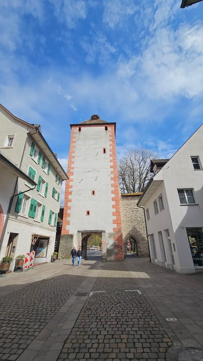 The image shows a street view in Rheinfelden, Switzerland. The "Oberturm" (Upper Tower) is prominently featured, a tall, white tower with orange brick detailing along the top and bottom edges. A pair of individuals are walking into the archway of the tower. The surrounding buildings have green shutters, and the street is paved with cobblestones. The sky above is blue with scattered clouds.