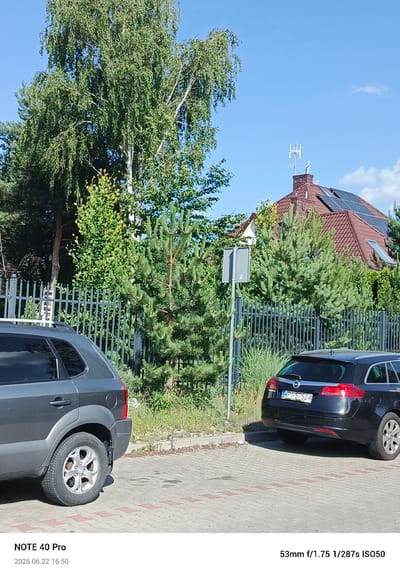 The image depicts an outdoor scene, likely in a residential area of Kamionka, Poland, on a sunny day. In the foreground, two cars are parked on a paved surface. The car on the left is a dark grey SUV, and the car on the right is a black station wagon. A metal fence runs behind the cars, separating the parking area from a grassy embankment and vegetation.

Behind the fence, there are various trees, including a large birch tree with white bark and lush green foliage, and several pine trees. A house with a red-tiled roof and solar panels is visible behind the trees. A television antenna is mounted on the chimney of the house. A street sign is also present near the fence. The sky is clear and blue with a few scattered clouds. The bottom of the image contains text indicating the camera model ("NOTE 40 Pro"), date and time ("2026.06.22 16:50"), and camera settings ("53mm f/1.75 1/287s ISO50"). There are no people visible in the image.