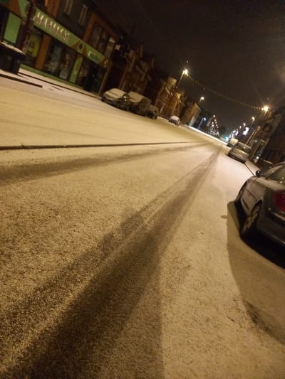A snow-covered street at night, illuminated by streetlights. Cars are parked along the side of the road. Dark tire tracks are visible in the snow, stretching down the road. Buildings line the street, some with lit windows. The image is slightly tilted, creating a sense of perspective.
