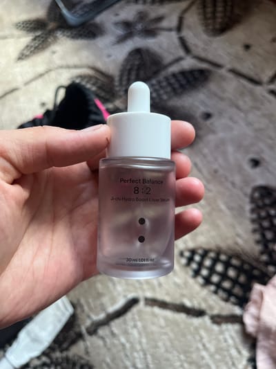 A person's hand holds a small, clear glass bottle with a white dropper lid. The bottle contains a pale pink serum. Text on the bottle reads "Perfect Balance 8:2 Ji-chi Hydro Boost Layer Serum 30 ml 1.01 fl. oz.". The background is a textured, neutral-colored surface, possibly a blanket or rug, with a black and white pattern. Some out-of-focus items, including what appears to be a black backpack with pink accents, are visible in the background. The lighting suggests an indoor setting during the daytime.