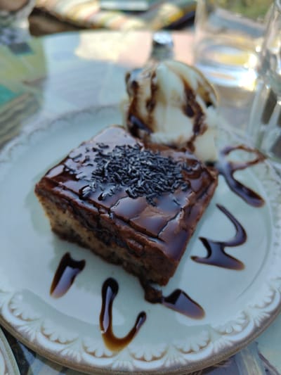 A square piece of chocolate cake, topped with chocolate frosting and dark sprinkles, sits on a light-colored, ornate plate. To the right of the cake is a scoop of vanilla ice cream drizzled with chocolate syrup. Chocolate syrup is also artfully drizzled around the cake and ice cream on the plate. The plate rests on a table with a patterned surface, possibly a tablecloth. In the background, a glass of water and other out-of-focus elements suggest an outdoor setting, likely a cafe or restaurant. The scene is set in Palaios Panteleimonas, Greece.
